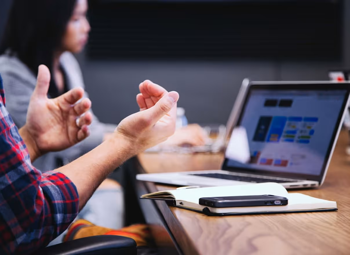 Person gesturing with hands near a laptop and notebook on a wooden table during a meeting.