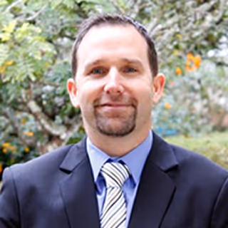 Man in a dark suit, blue shirt, and striped tie, smiling outdoors with trees in the background.