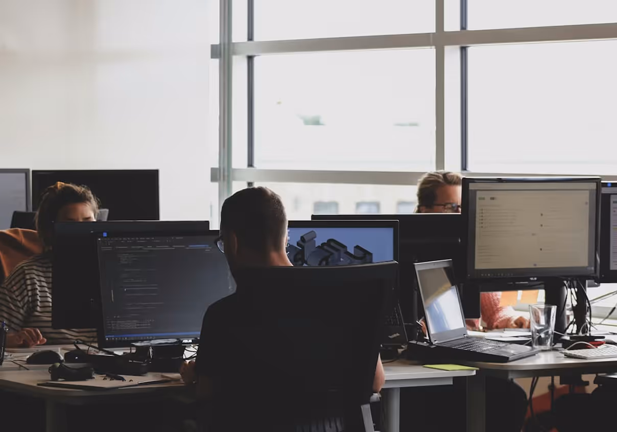 People working at desks with multiple monitors and laptops in a modern office with large windows.