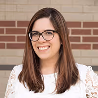 Smiling woman with long brown hair and glasses wearing a white lace top standing in front of a brick wall.