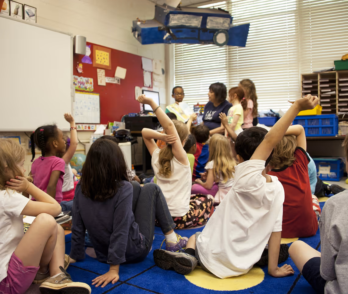 Group of children sitting on a colorful rug in a classroom, some raising their hands while a teacher reads a book to them.