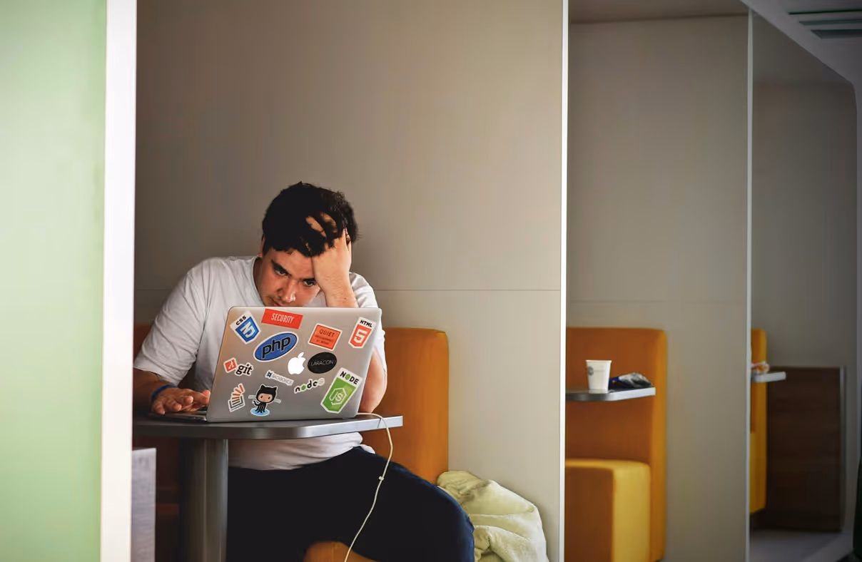 Person sitting at a table in a booth with a laptop covered in programming and tech stickers, looking stressed and holding their head.