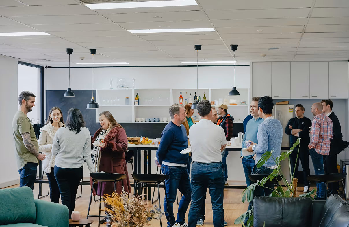 Group of people standing and chatting in a modern kitchen or break room with white cabinets and hanging pendant lights.