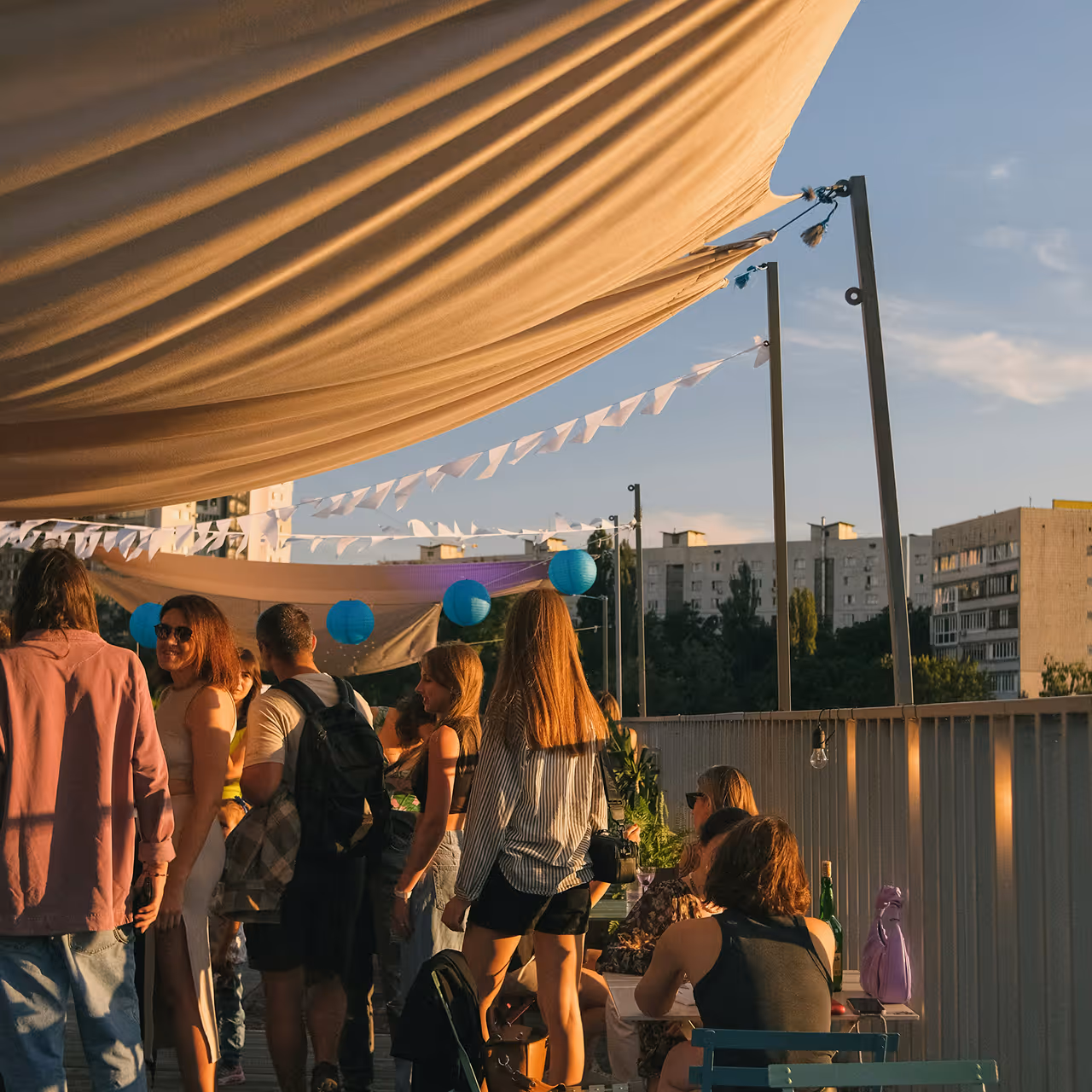 Group of people socializing under large fabric awnings with blue paper lanterns during a sunny outdoor rooftop gathering.