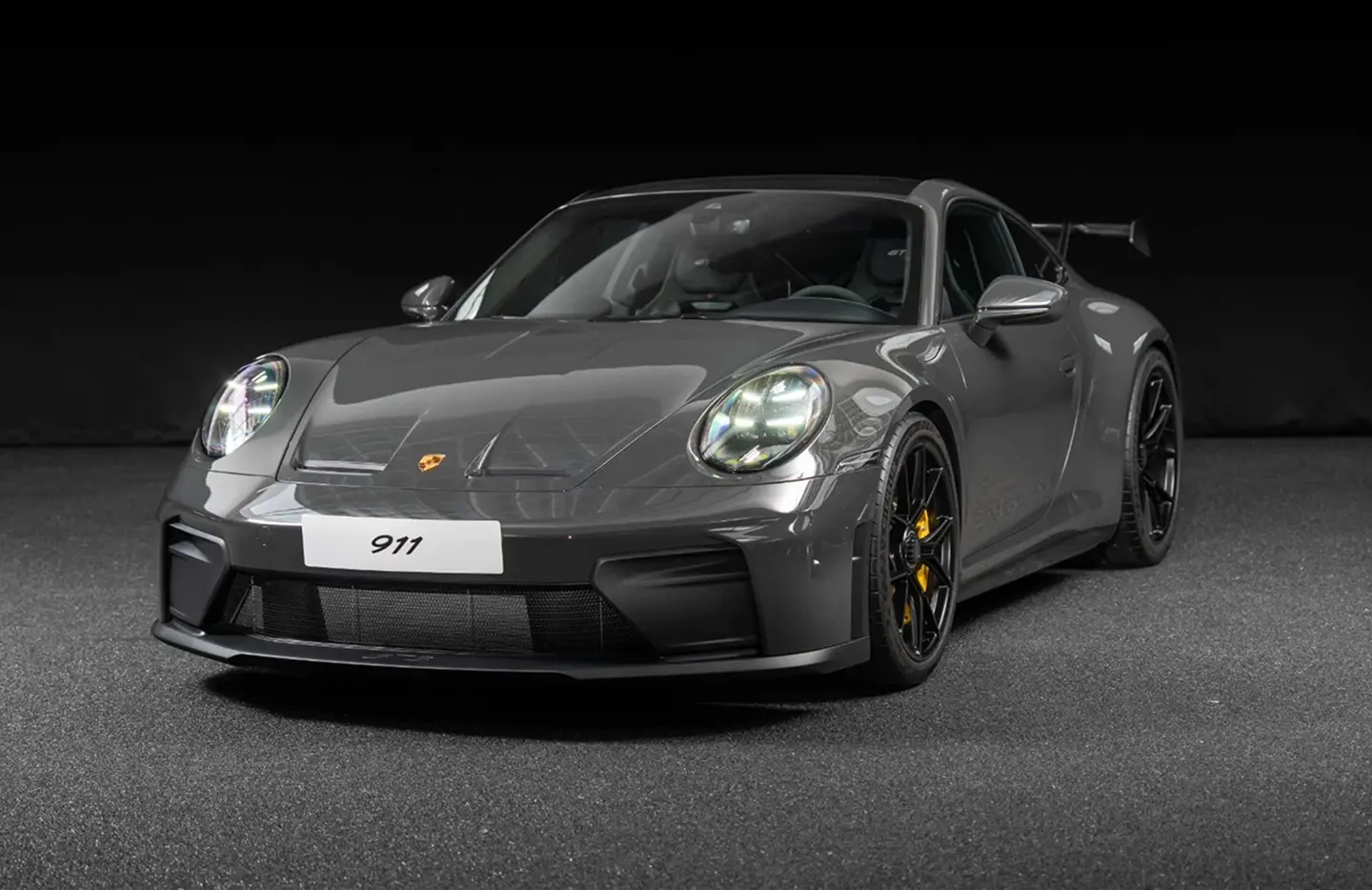 Front angled view of a gray Porsche 911 sports car with illuminated headlights and black wheels against a dark background.
