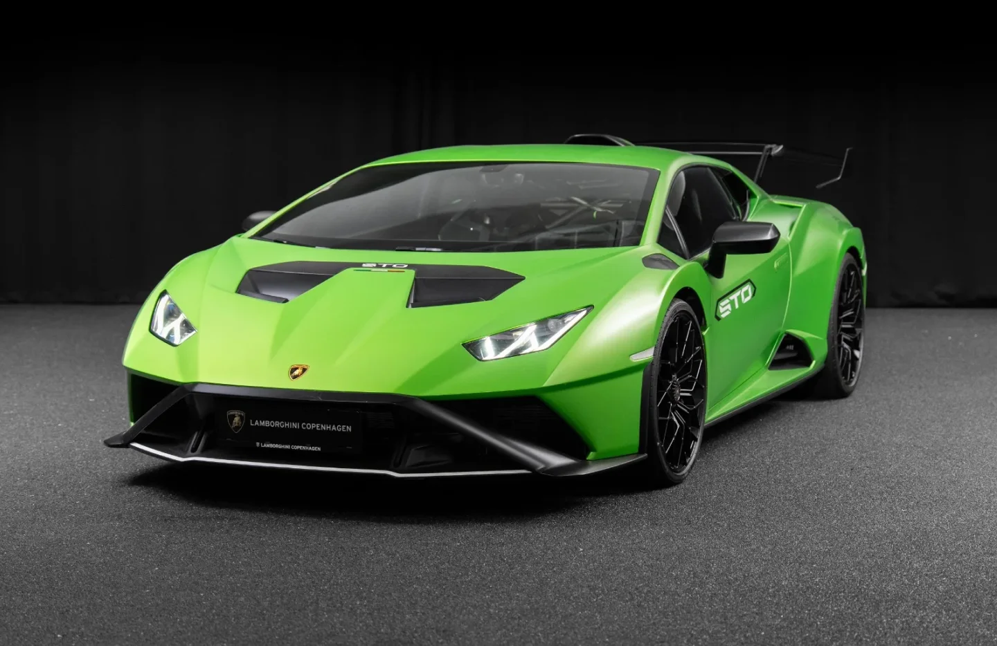 Bright green Lamborghini Huracán STO sports car with black accents and aerodynamic features, parked on a dark textured floor against a black background.