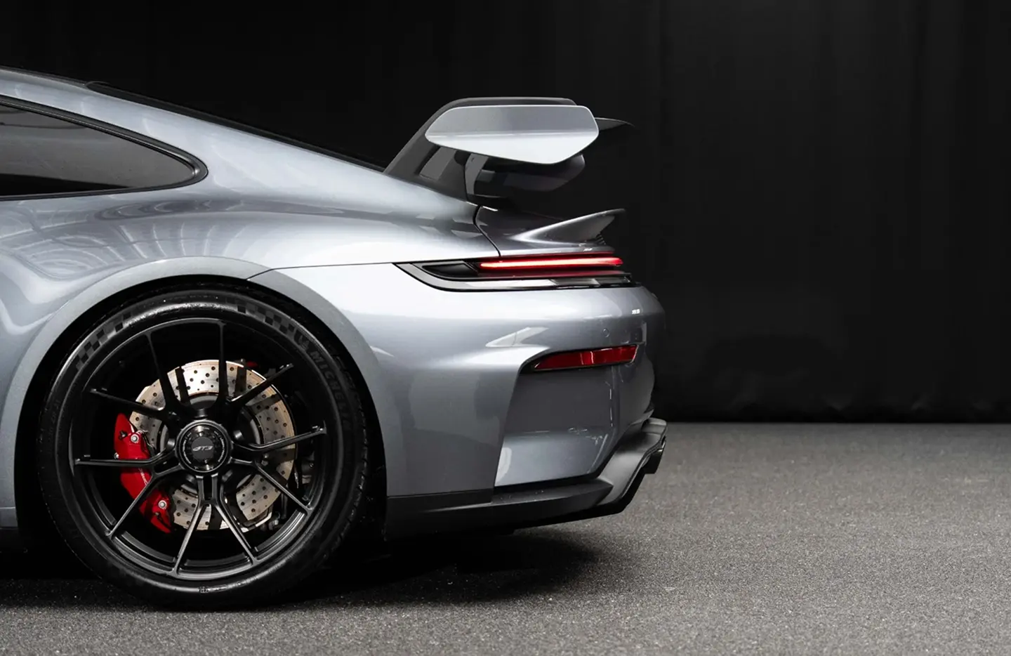 Rear side view of a silver sports car with black alloy wheels, red brake calipers, and a raised spoiler against a black background.