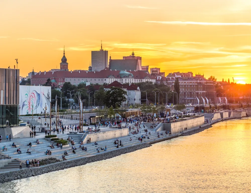 People relaxing on riverside steps at sunset with Warsaw's Old Town skyline in the background.