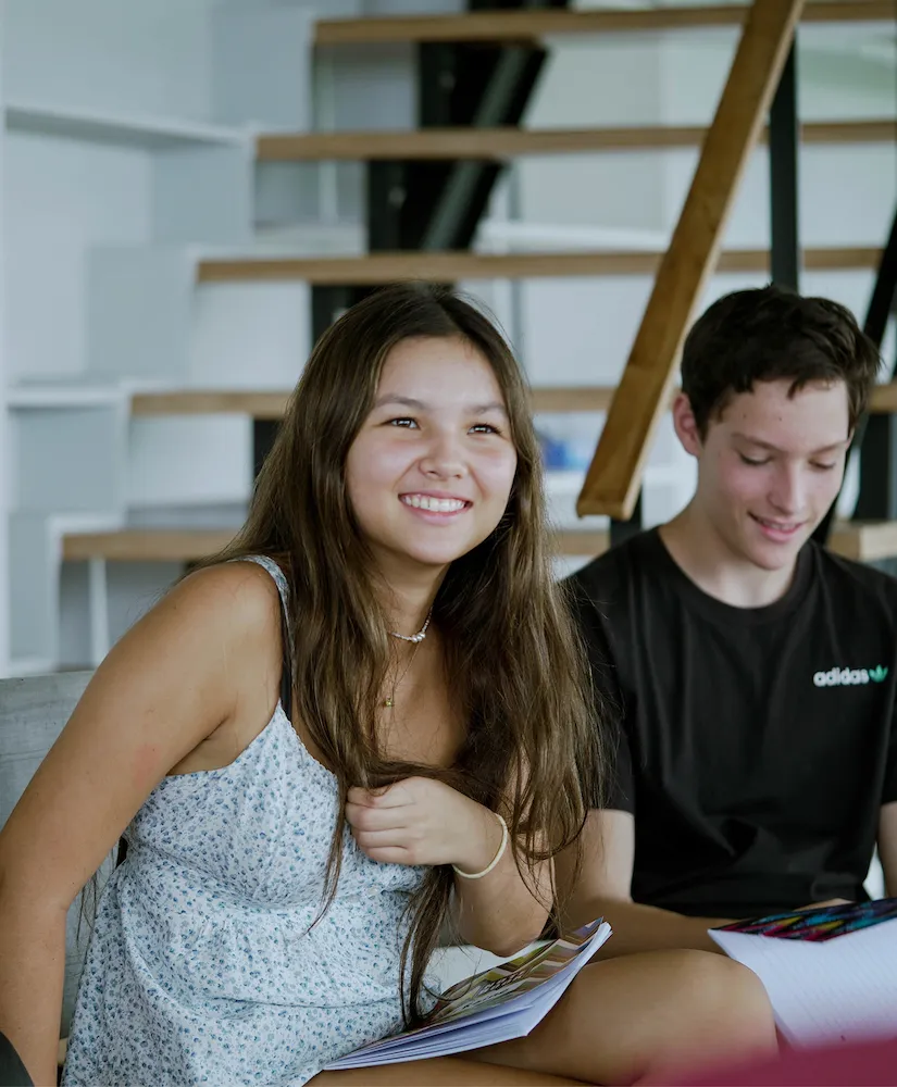 Two young people smiling indoors, holding notebooks, sitting near a wooden staircase.