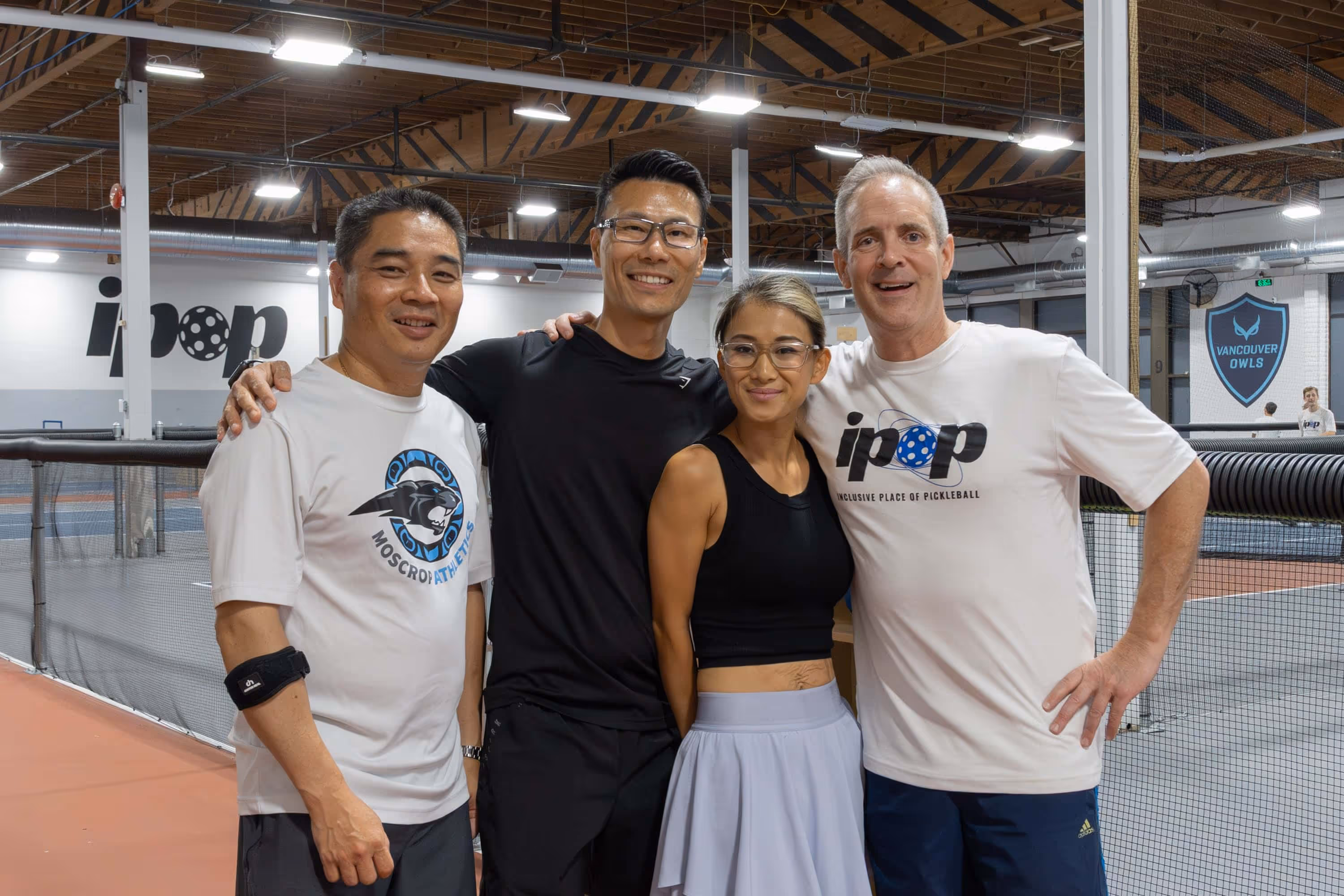Four people standing together smiling inside an indoor pickleball court with nets and signage visible.