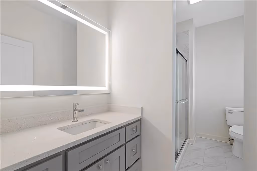 Modern bathroom with gray vanity, illuminated mirror, glass shower door, and white toilet.