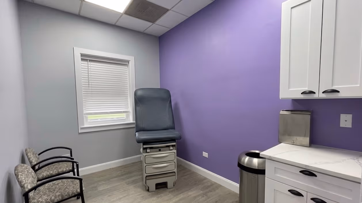Medical exam room with gray exam chair, two patterned waiting chairs, purple wall, and white cabinets.