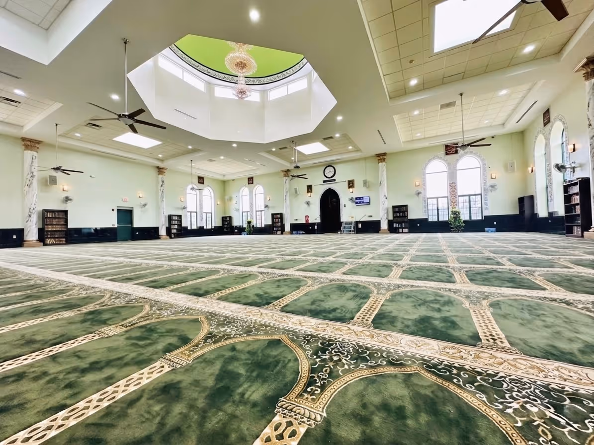Spacious mosque interior with green patterned carpet, arched windows, chandeliers, and ceiling fans.