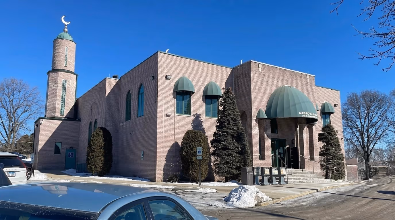 Brick Islamic center with green domes and crescent, parked cars, snow patches, and clear blue sky.