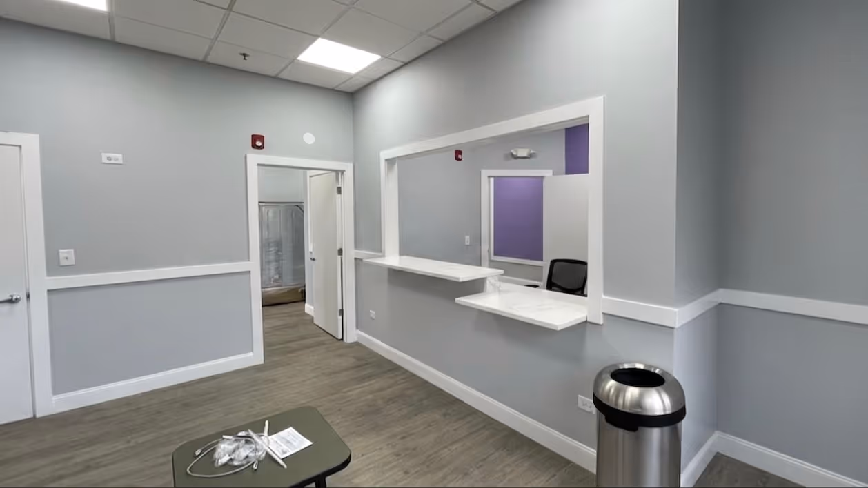 Medical office reception area with a counter window, chair, trash can, and open door to a room with shower.