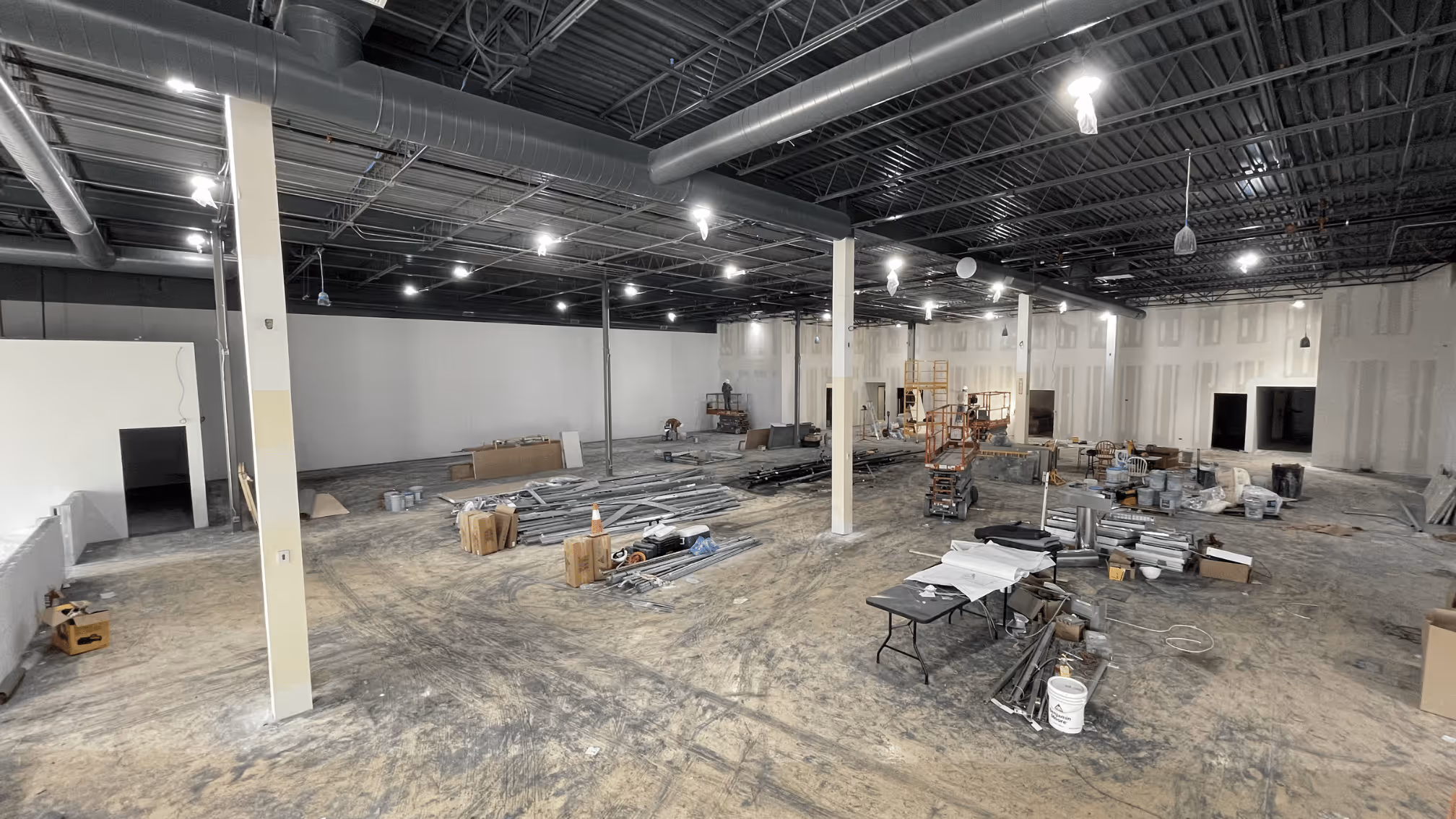 Large indoor construction site with metal beams, equipment, and workers in an unfinished building.