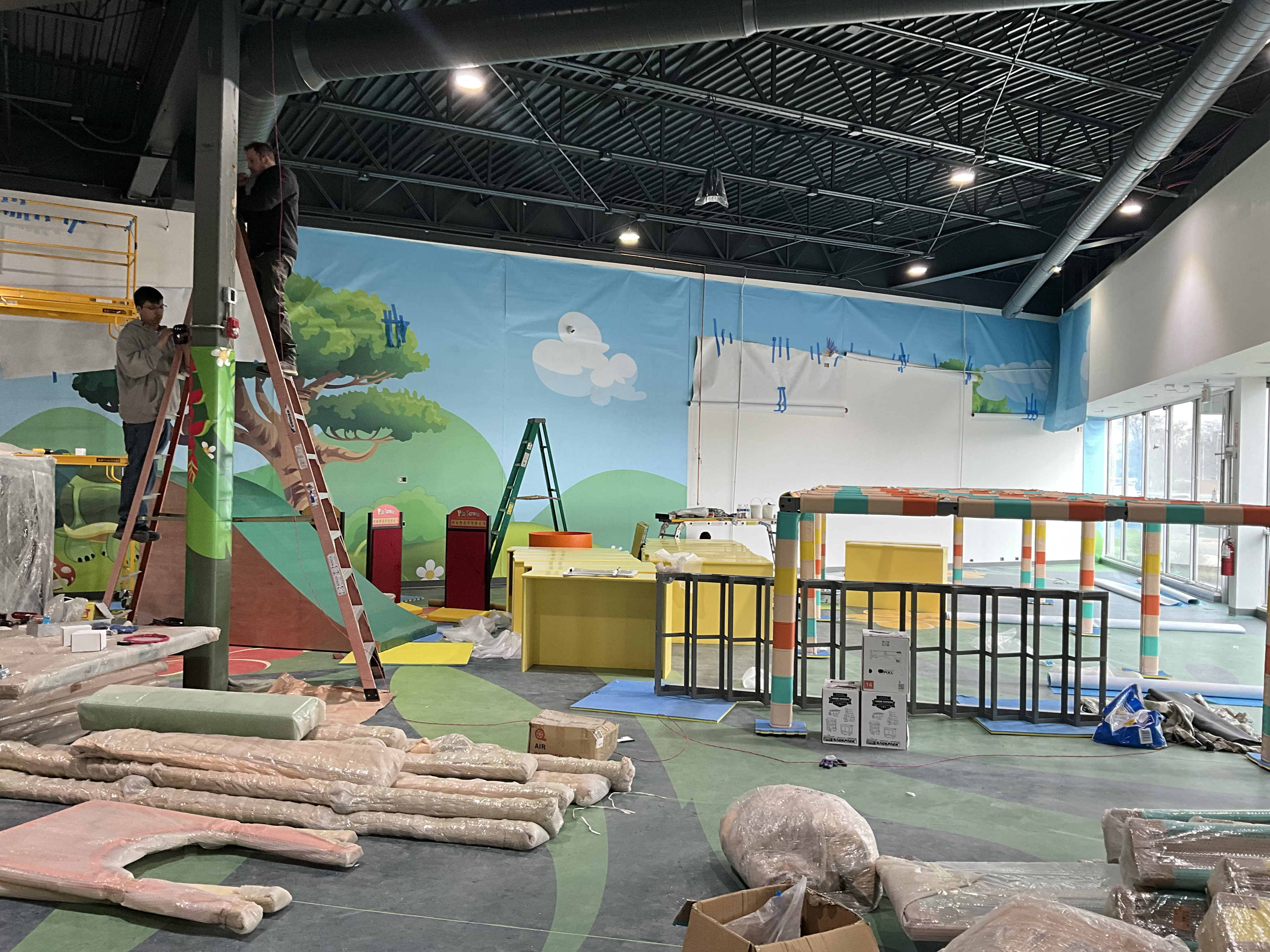 Indoor children's play area under construction with colorful floors, murals, and workers on ladders.