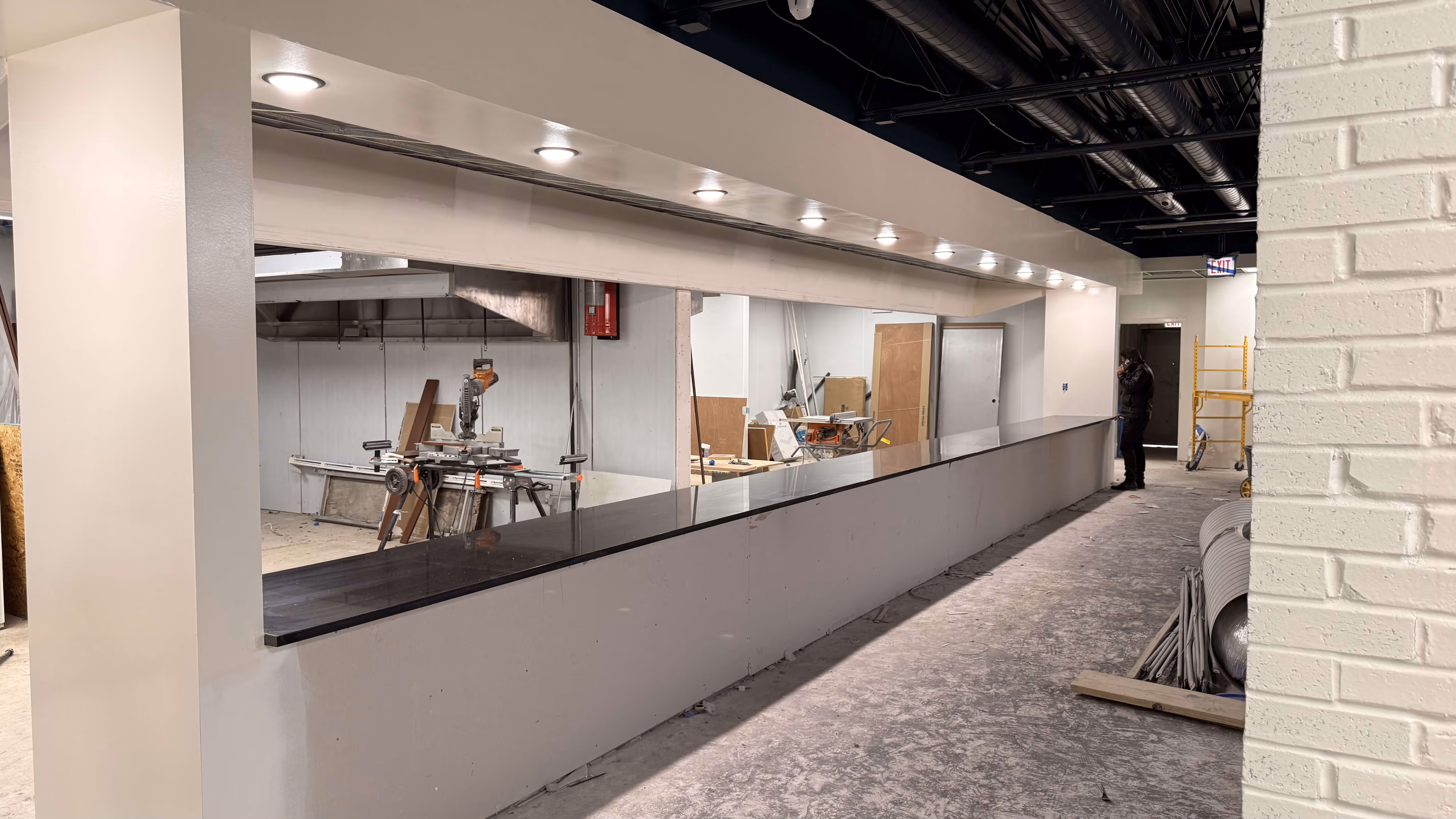 Long counter with black countertop in an unfinished room with construction tools and person on phone.