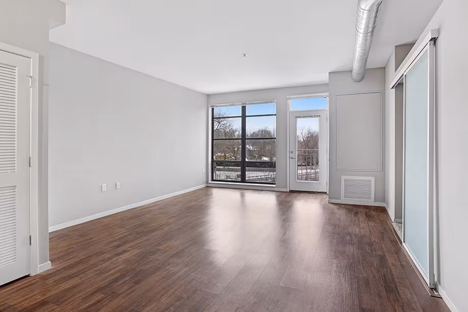 Empty modern apartment living room with wood floors, large window, glass door, and exposed duct.