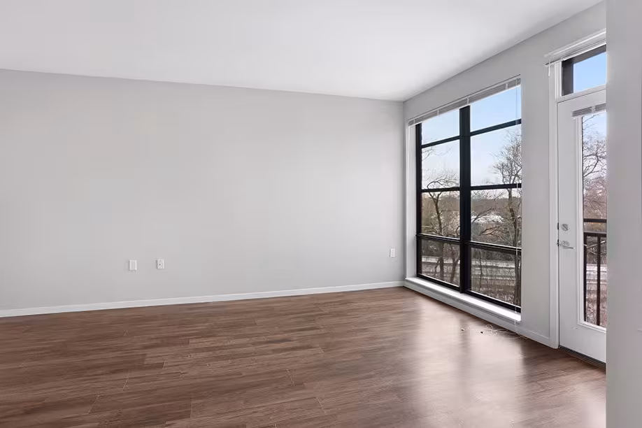 Empty room with wooden floor, large black-framed window, and glass door leading to balcony.