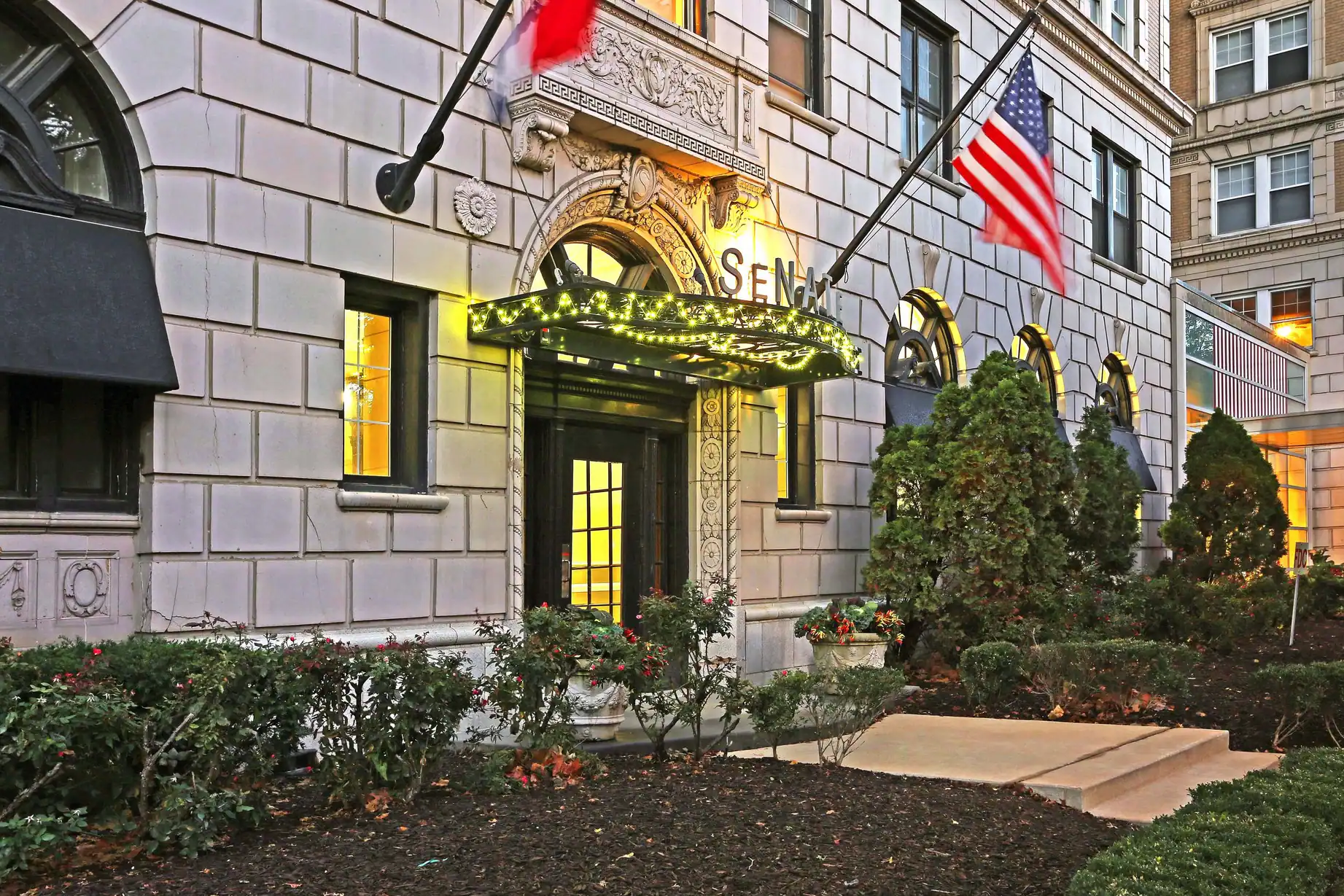 Entrance to a classical building with illuminated canopy, American flag, and landscaping with shrubs.