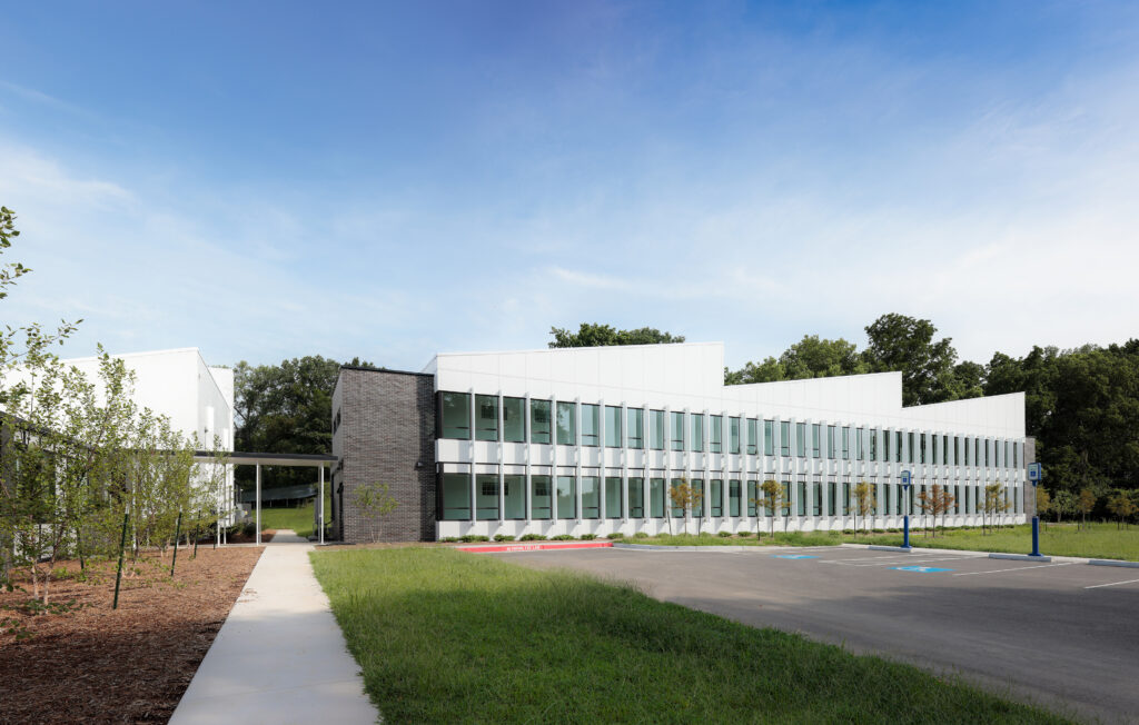 Modern two-story building with large windows, adjacent sidewalk, parking spaces, and surrounding trees.