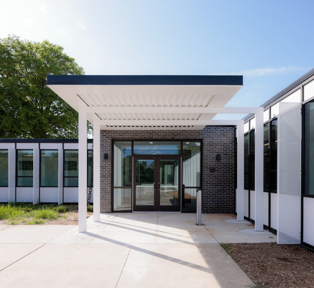 Modern building entrance with white pergola, glass double doors, and brick accent wall under clear sky.