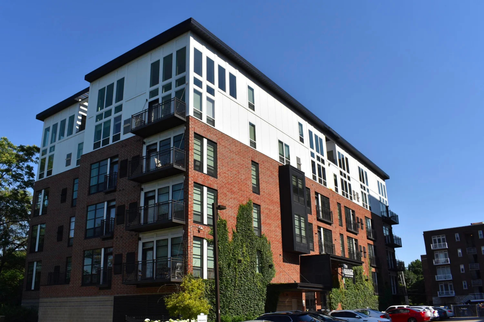 Four-story apartment building with brick and white panels, balconies, and ivy on the walls under clear blue sky.