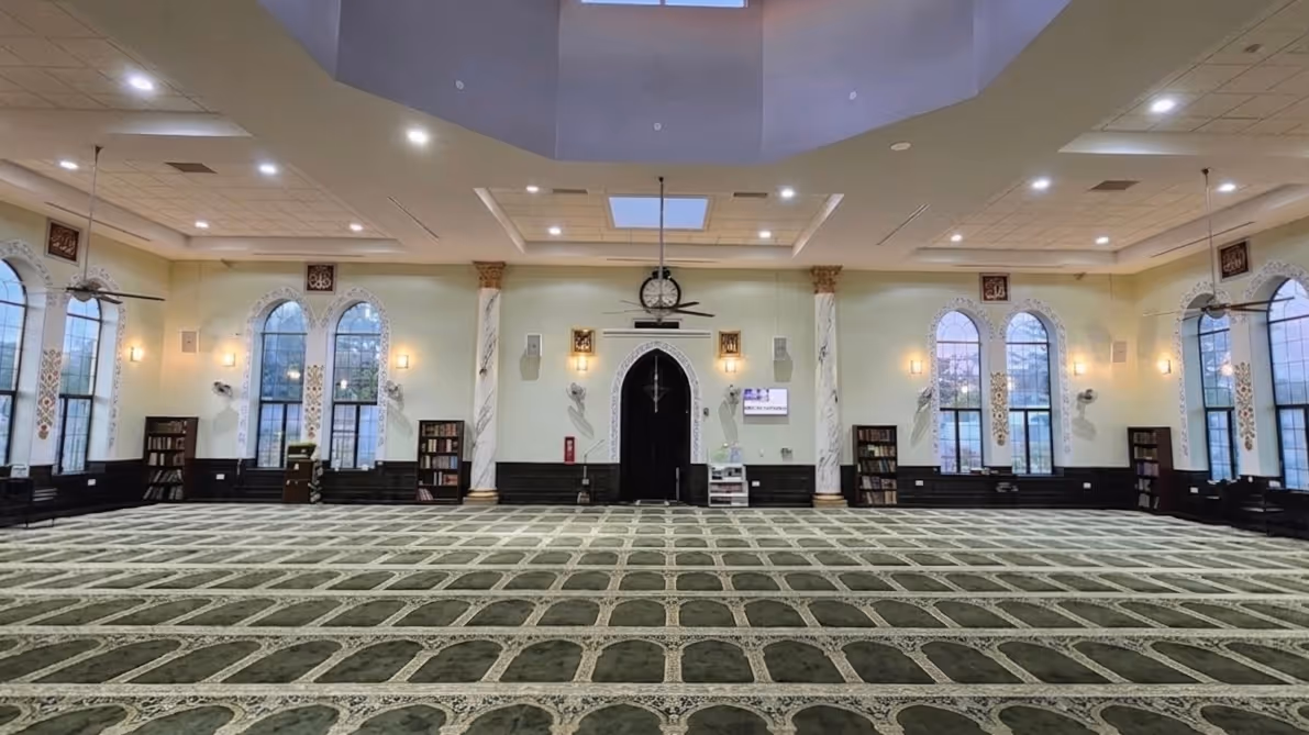 Spacious mosque interior with green patterned carpet, arched windows, chandeliers, and ceiling fans.