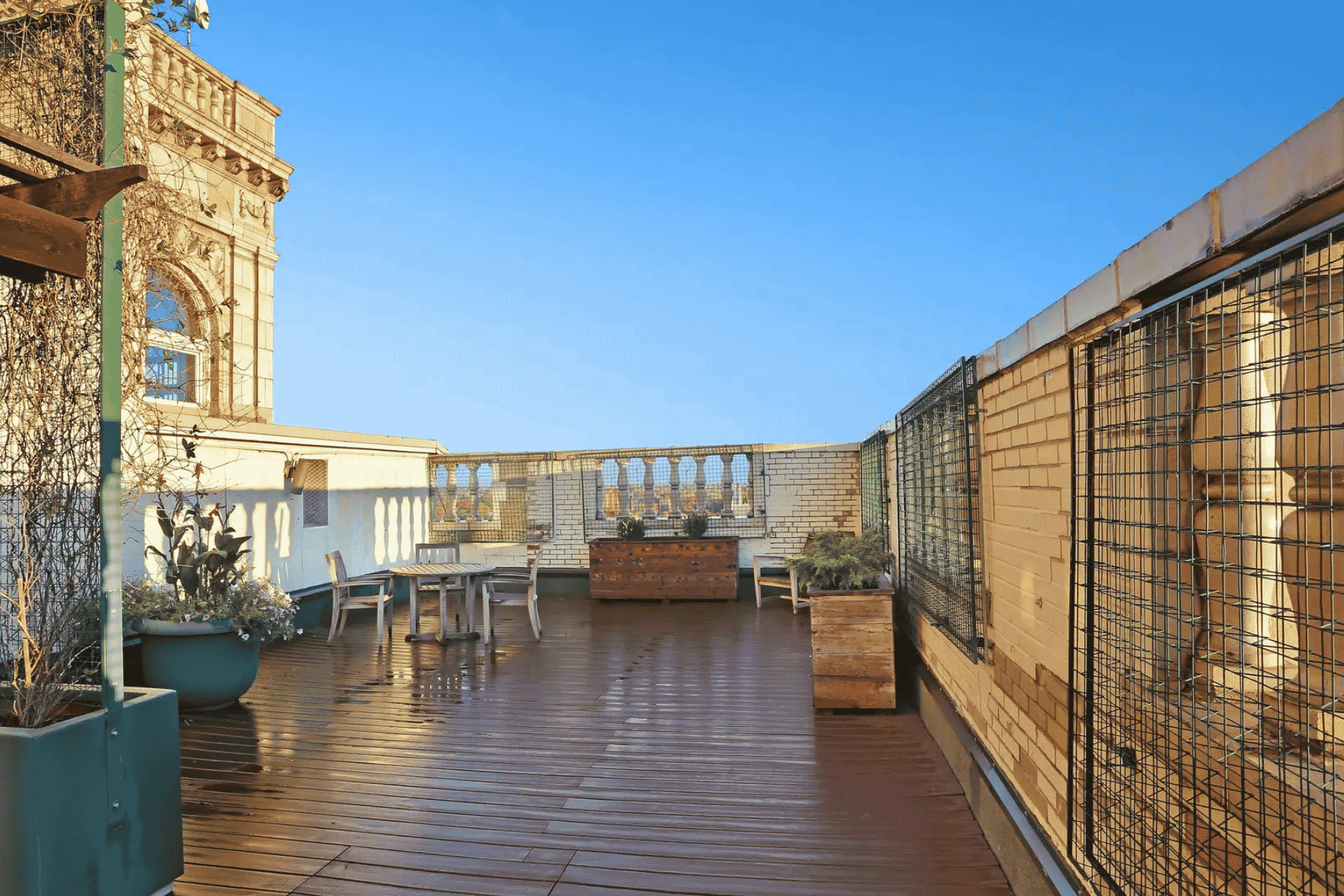 Spacious rooftop terrace with wooden deck, chairs, tables, and potted plants under a clear blue sky.