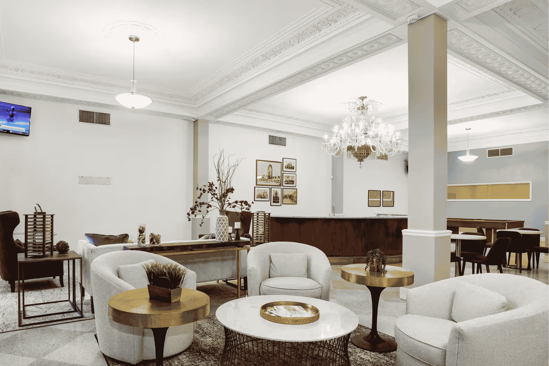 Elegant lounge area with gray armchairs, gold tables, chandelier, and wooden reception desk in bright room.