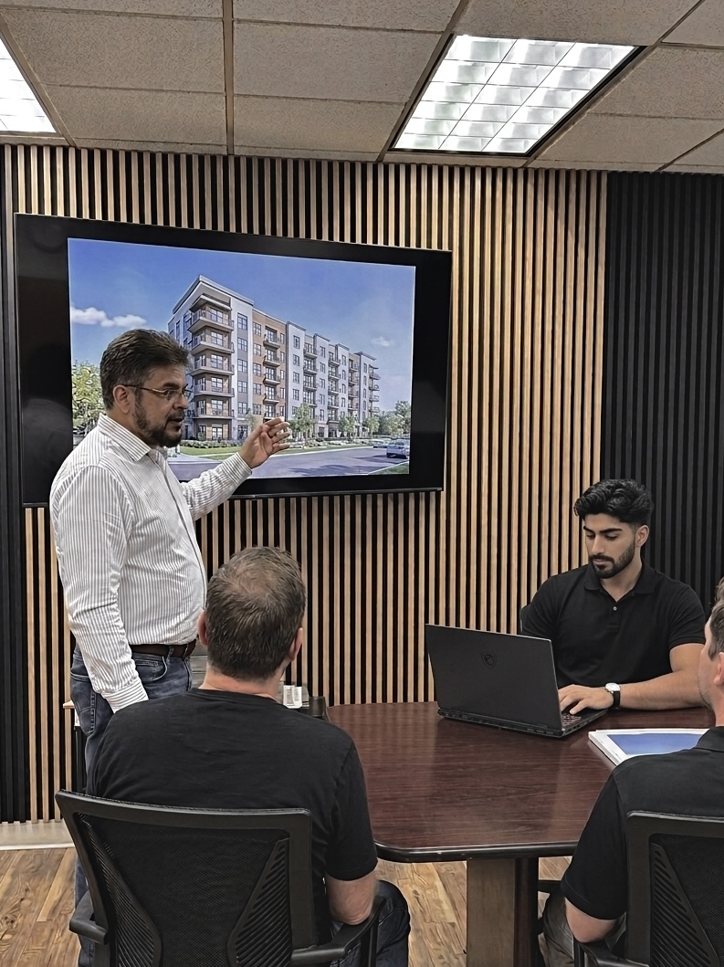 Man stands pointing to a building floor plan on a screen as four others sit at a conference table.