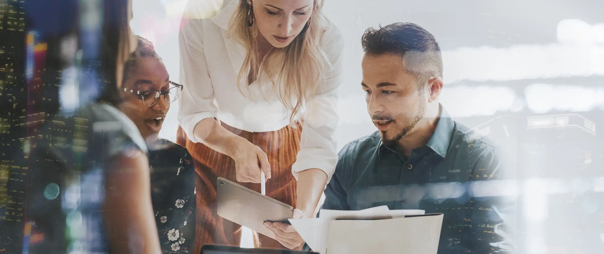 Four young business professionals in a modern-looking office with three of them glancing at a tablet.