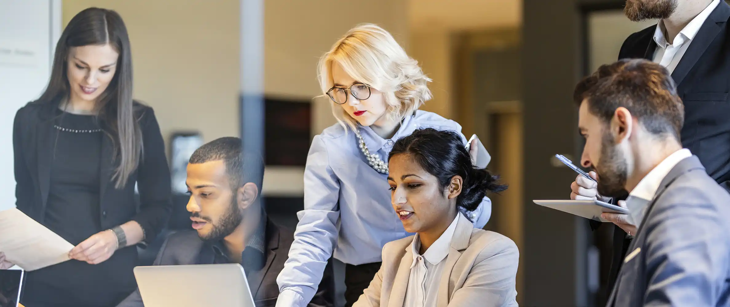 A group of businesspeople in an office work together at an open-concept desk. Three are reviewing something on a laptop.