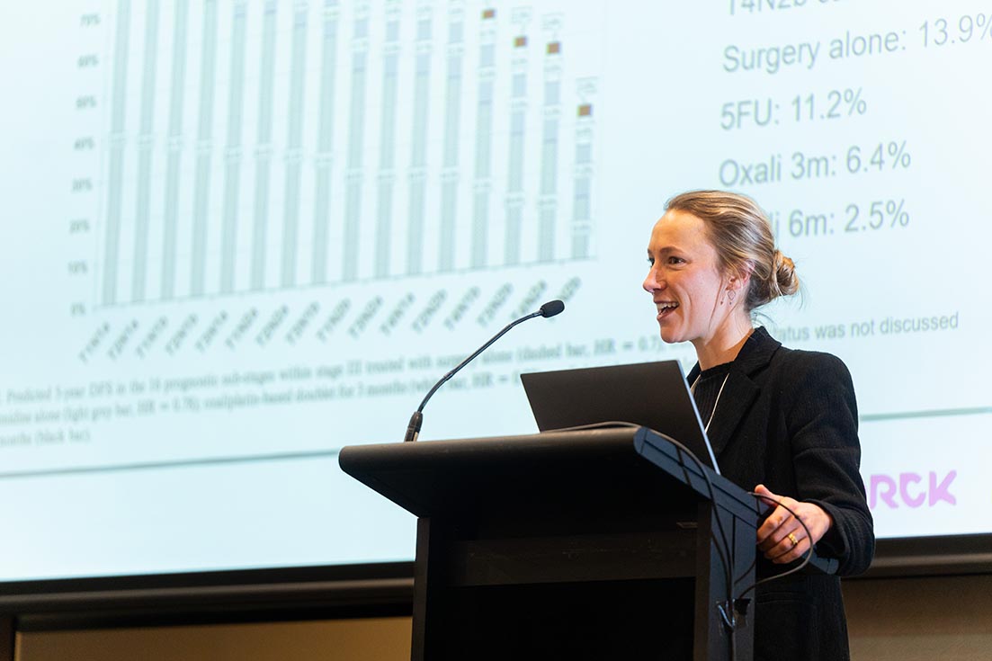 A woman speaking at a lectern during a medical presentation. Behind her, a projector screen shows a bar chart and statistical data.