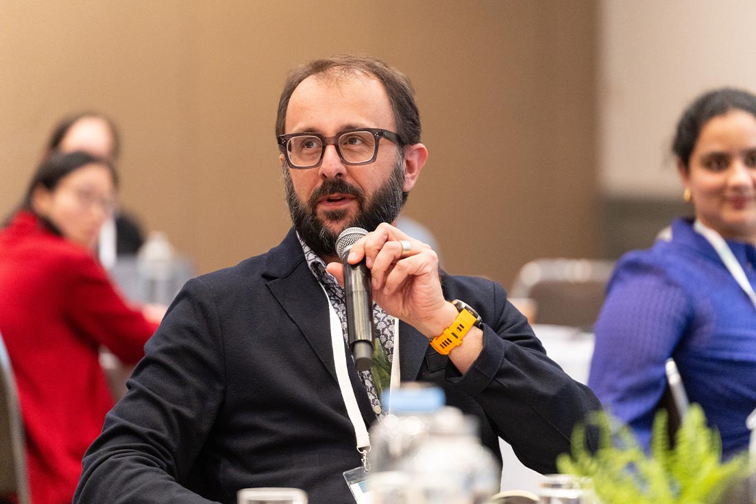 A man with a beard and glasses speaking into a microphone at a professional conference or seminar table.