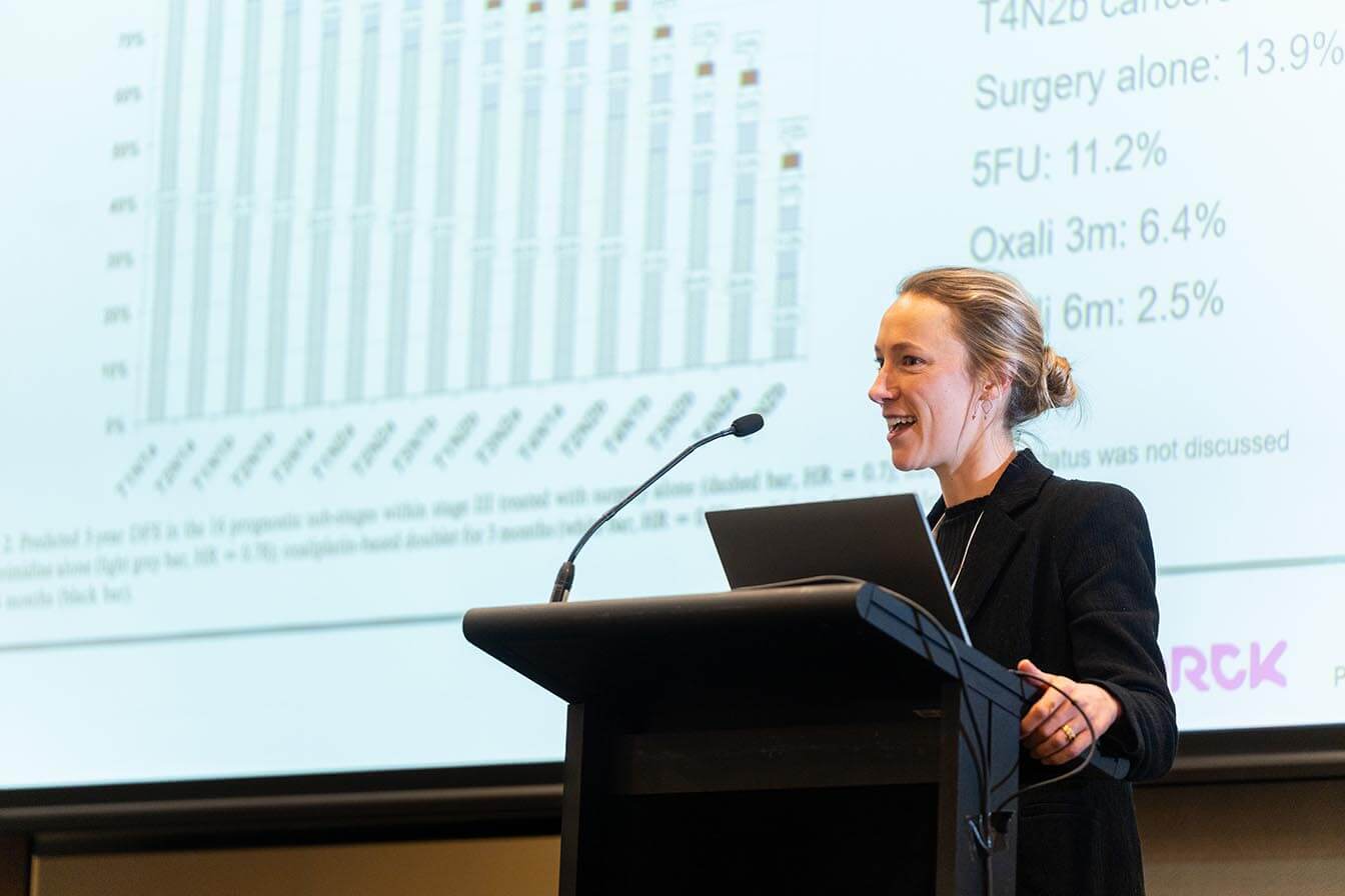 A female researcher presenting cancer survival data from a podium, with a bar chart projected on the screen behind her.