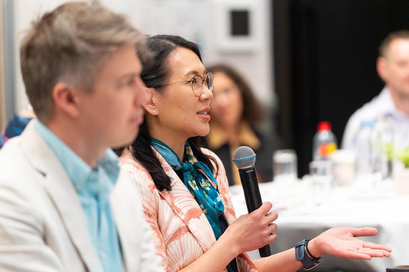 A woman in a floral blazer holding a microphone and explaining a point during a panel discussion at a research event.