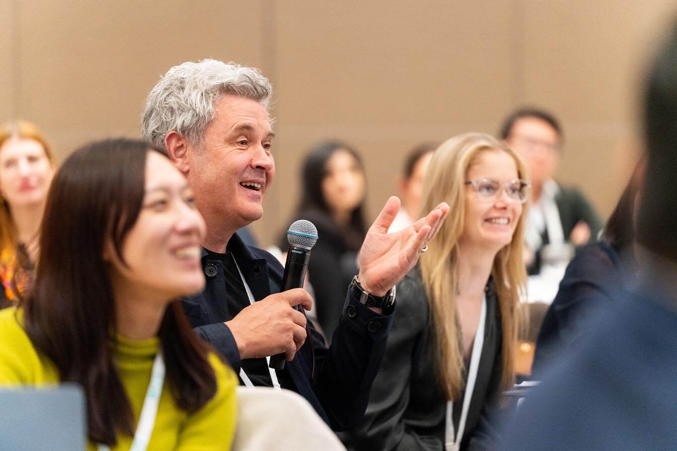 A man with grey hair gesturing with his hand while asking a question during a Q&A session at a conference.
