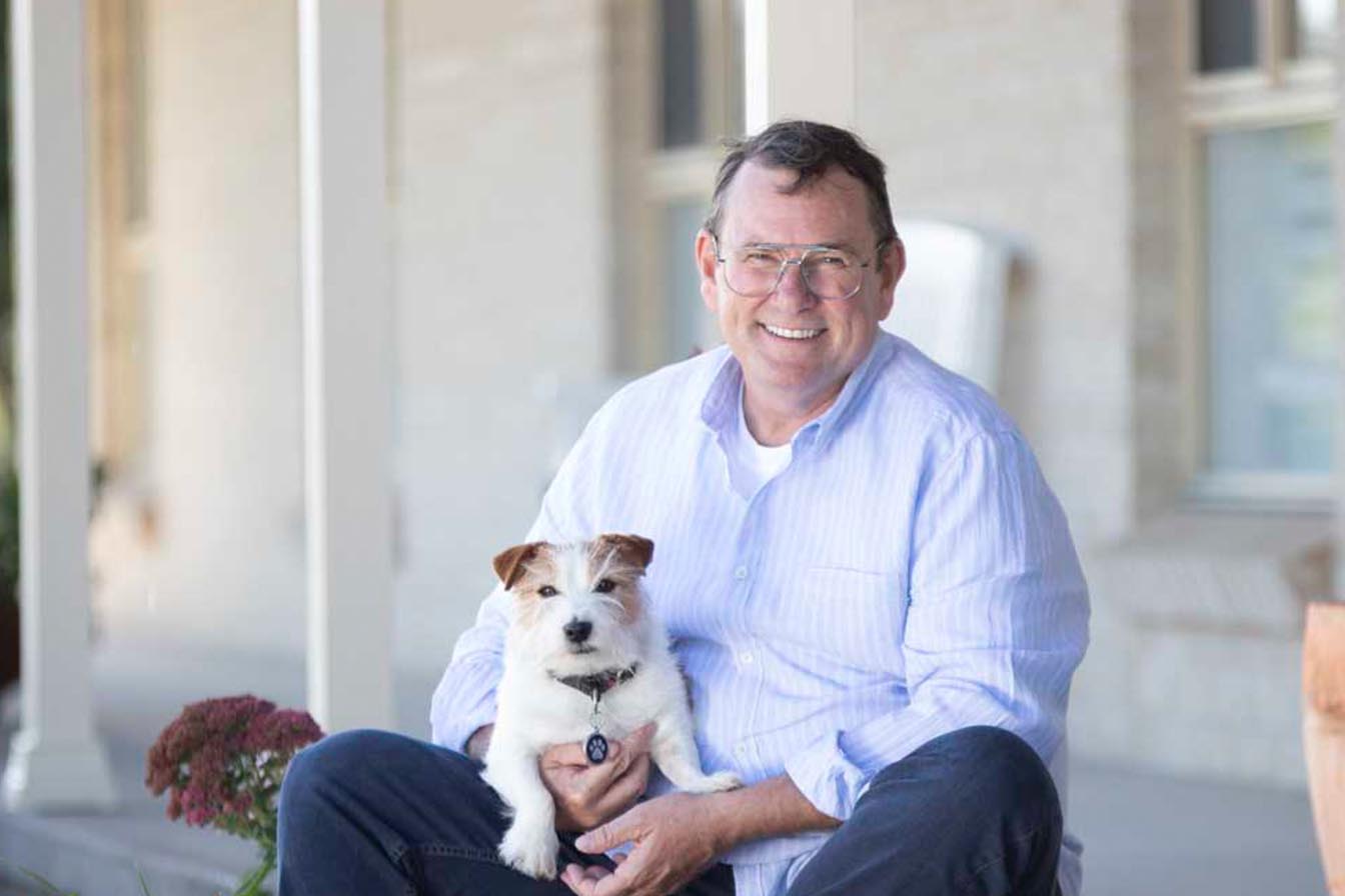 A smiling man in a light blue shirt and glasses sitting outdoors on a porch holding a small white and brown dog.