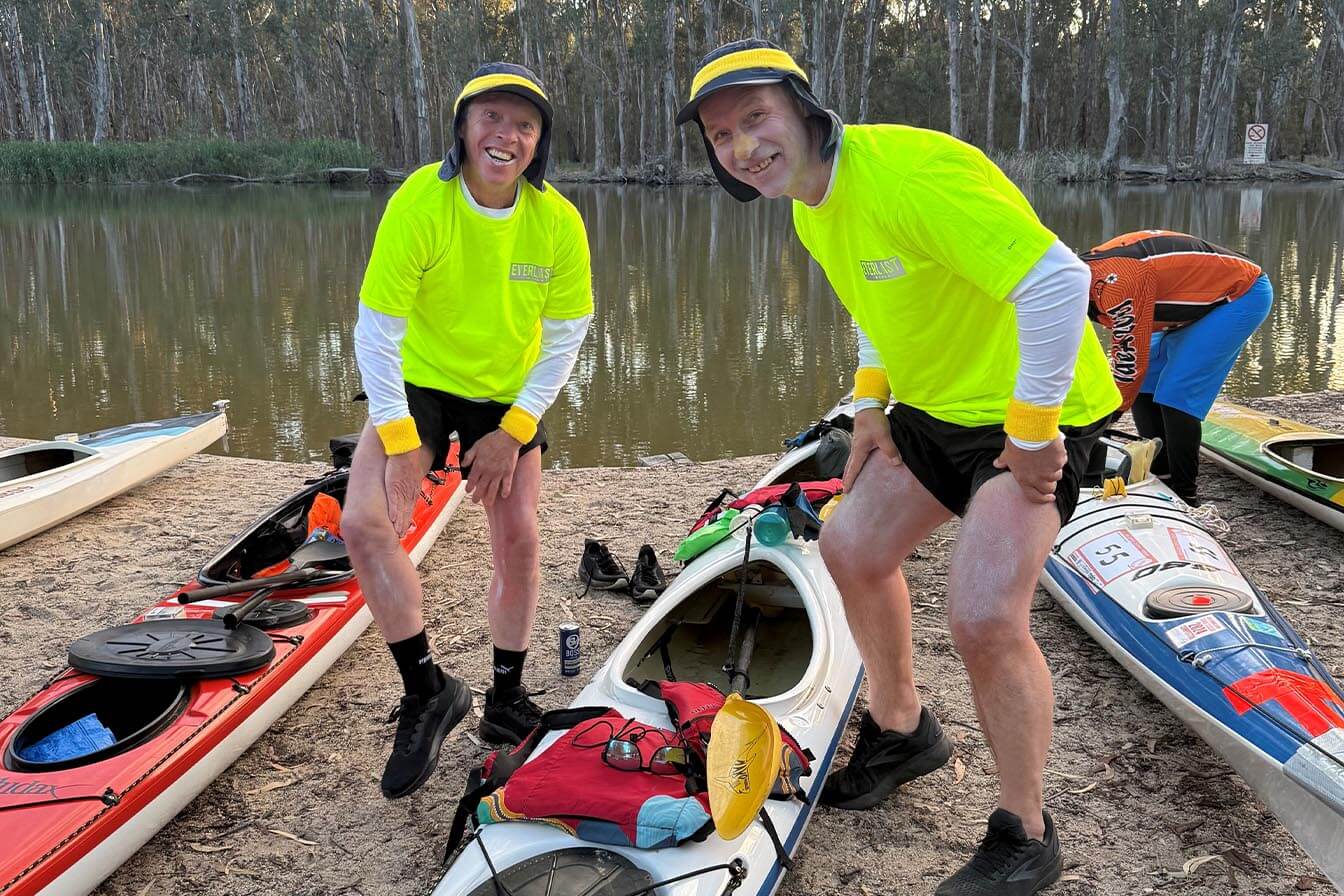 Two men in bright neon yellow shirts and sun hats smiling next to their kayaks on a riverbank.
