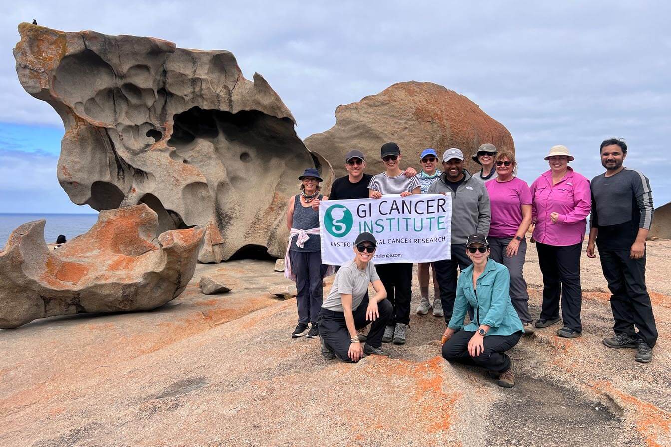 A group of travellers posing with a GI Cancer Institute banner in front of the Remarkable Rocks on Kangaroo Island.