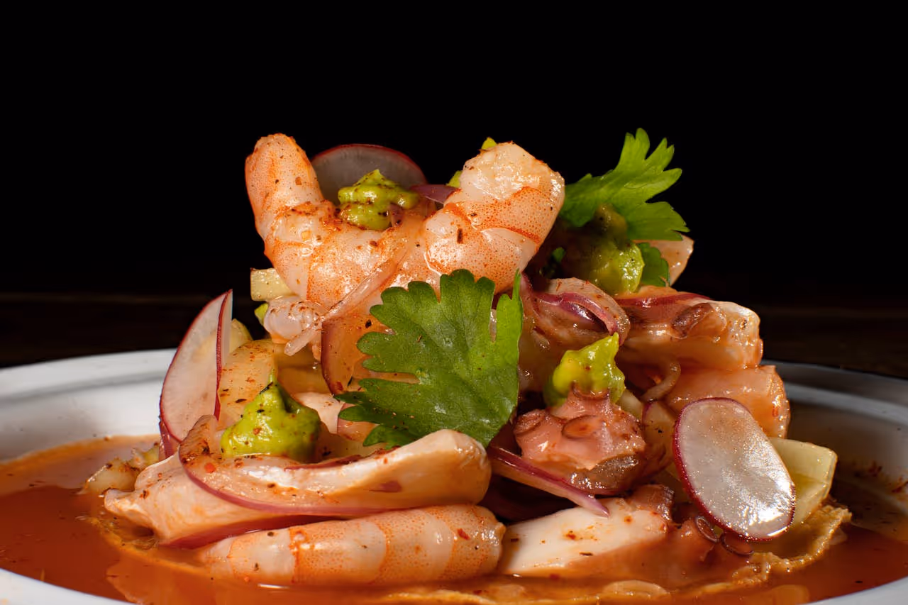 Plate of shrimp ceviche garnished with cilantro, radish slices, and avocado sauce in a red sauce.