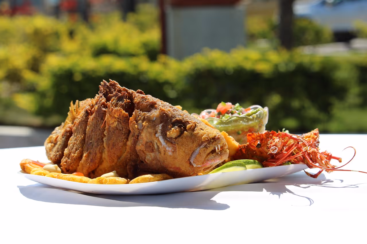 Fried whole fish served on a white plate with lime slices, lobster, fried plantains, and a cup of guacamole salsa on an outdoor table.