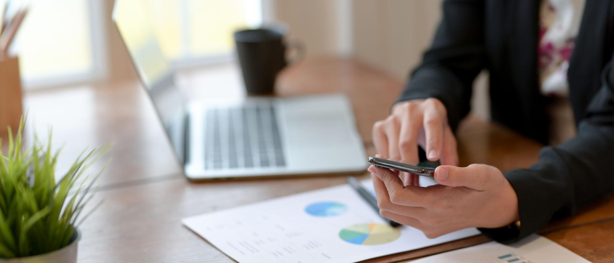 Person using a smartphone over a desk with a laptop, coffee cup, potted plant, and printed charts.