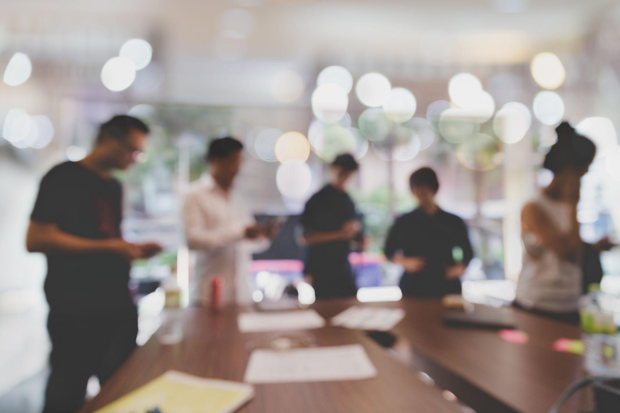 Blurry image of five people standing around a table with papers and drinks in a bright office.