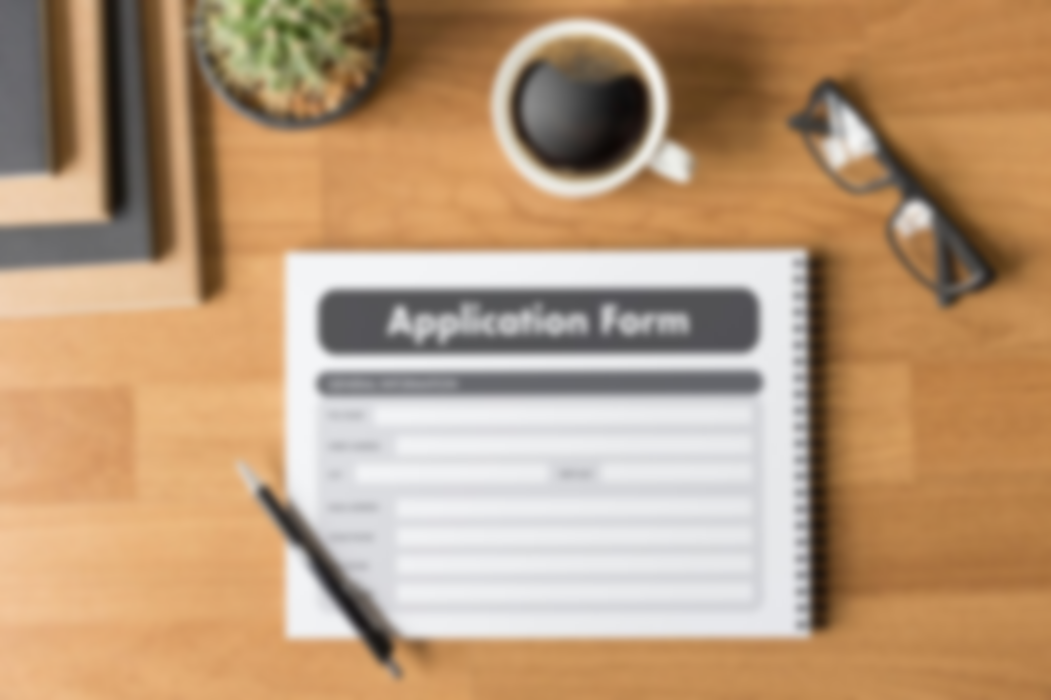 Top view of a desk with an application form, a pen, a cup of coffee, black-framed glasses, a succulent, and stacked notebooks.