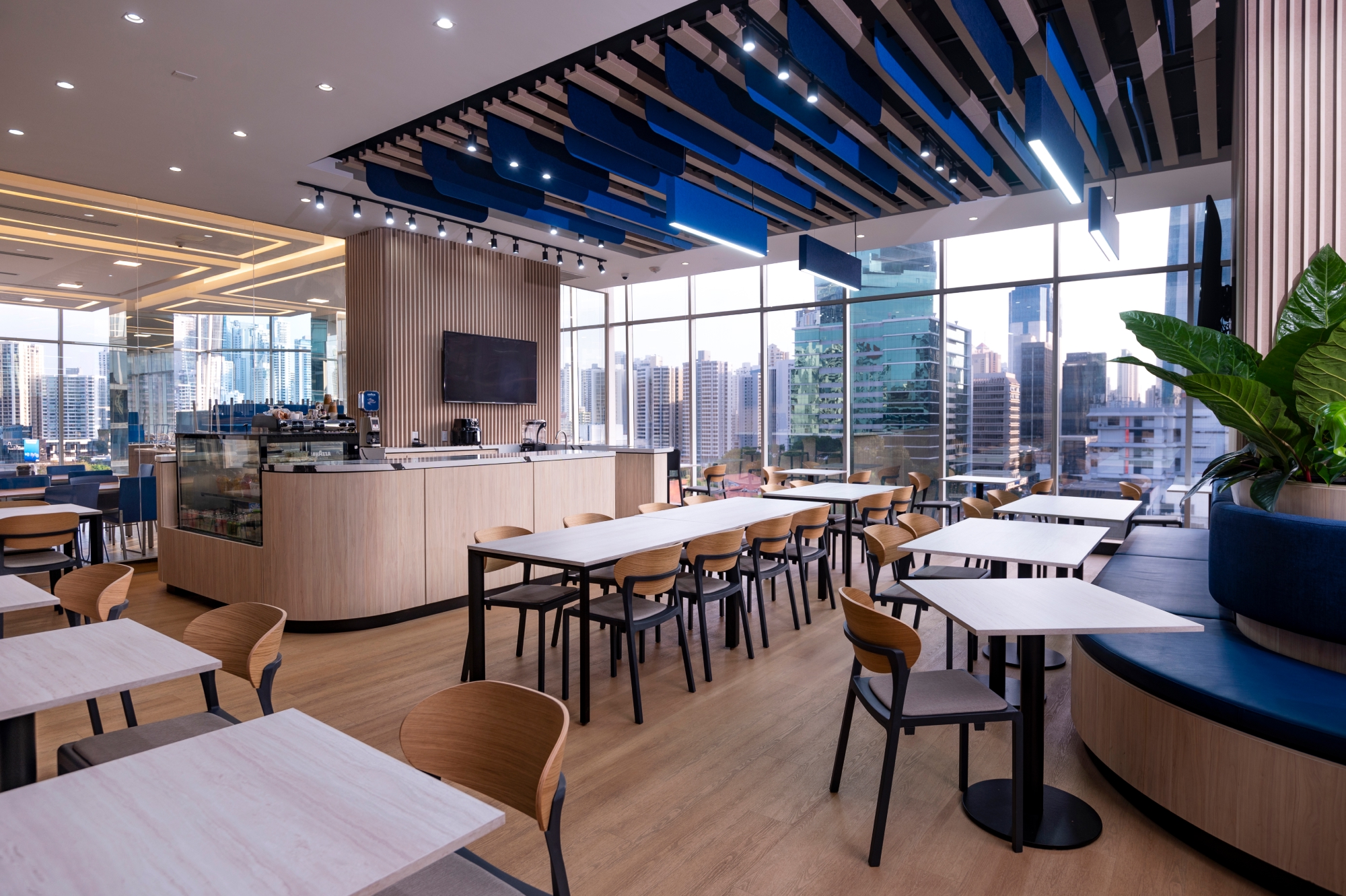 Modern office cafeteria with light wood tables, chairs, blue cushioned seating, and large windows showing a city skyline.