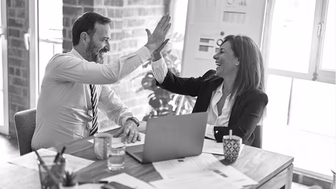 Success with CAPDA. Man and woman in business attire giving each other a high five at an office desk with a laptop and papers.