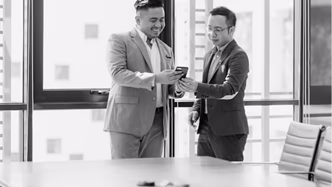 Success with CAPDA. Two men in business attire smiling and looking at a smartphone in an office with large windows.